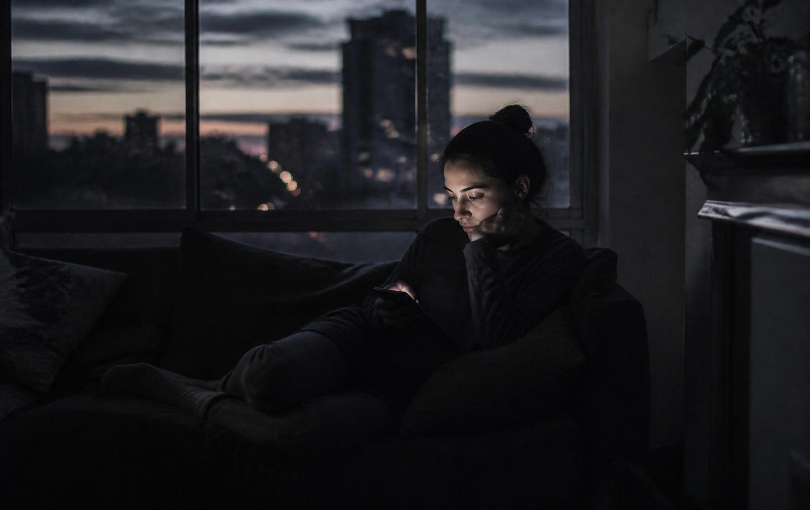 A person sitting alone on a couch at dusk, lit only by a phone screen